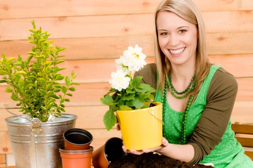 A young woman with blonde hair and a bright smile sits in an outdoor garden space, holding a yellow flowerpot containing a healthy, blossoming white flowering plant. She is wearing a green blouse with a matching bead necklace and a brown cardigan. Next to her on a wooden table are multiple plant pots, including a metallic container with a small, bushy green shrub, a larger black pot, and an empty terracotta pot. The background features a wooden fence or wall, contributing to a warm, natural outdoor setting. The garden area appears well-maintained, with a variety of potted plants and gardening tools visible, suggesting outdoor gardening activity. Natural daylight illuminates the scene, highlighting the vibrant green foliage and the bright white flowers, creating a cheerful and inviting atmosphere consistent with professional gardening or landscaping services such as those offered by Lawn Mowing Battersea in the Battersea area.