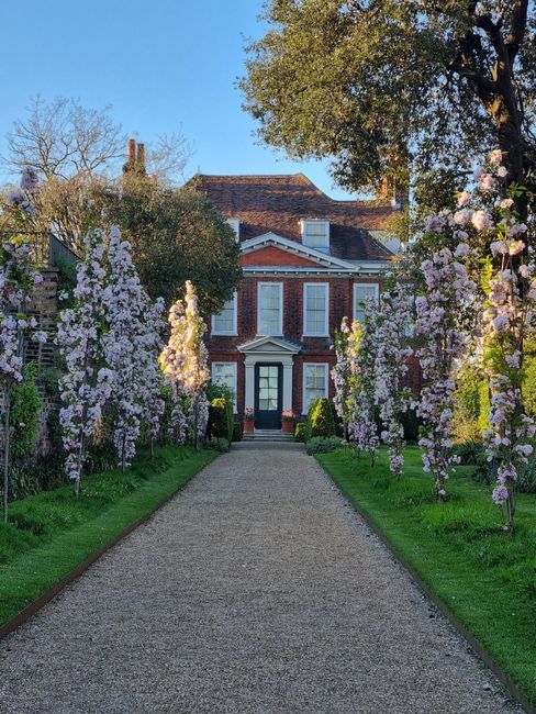 A symmetrical garden pathway with a light-colored gravel surface leads through a well-maintained landscape, flanked by neatly trimmed grass and a series of tall, slender cypress trees on both sides. O