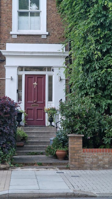 Close-up view of lavender-colored flowering plants with slender, green, slightly fuzzy leaves in the foreground, growing in a narrow front garden area. Behind the plants, there is a Victorian-style te